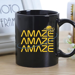 May include: Black ceramic mug with the word "AMAZE" in yellow, repeated three times, with a blue swirl and a yellow crab graphic. The mug has a curved handle.