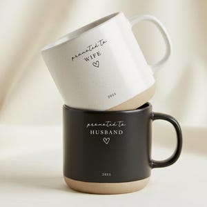 May include: Two ceramic mugs, one white and one black, stacked on top of each other. The white mug has the text "promoted to WIFE" and the black mug has the text "promoted to HUSBAND". Both mugs have a heart symbol and the year 2025.