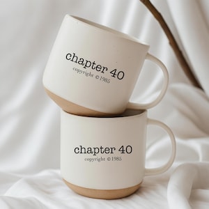 May include: Two white ceramic mugs stacked on top of each other. Each mug has a tan base and a curved handle. The text "chapter 40 copyright © 1985" is printed in black on the front of each mug.