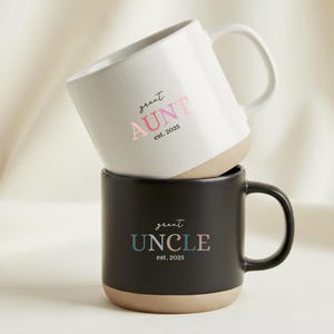 May include: Two ceramic mugs, one white and one black, stacked. The white mug has the text "great AUNT est. 2025" in pink. The black mug has the text "great UNCLE est. 2025" in blue and green. Both mugs have a tan base.