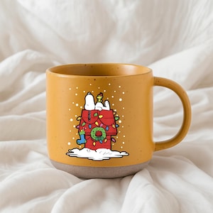 May include: Mustard yellow ceramic mug featuring a festive illustration of Snoopy's doghouse decorated for Christmas. The red doghouse is adorned with Christmas lights, a wreath, and a stocking. Snoopy and Woodstock are on top of the doghouse.