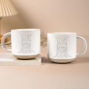Op de afbeelding: Twee witte keramische mokken met een boomontwerp en een hartvormig embleem. Het embleem bevat initialen en een datum. De mokken hebben een beige basis en een gebogen handvat. De mokken staan op een wit oppervlak.