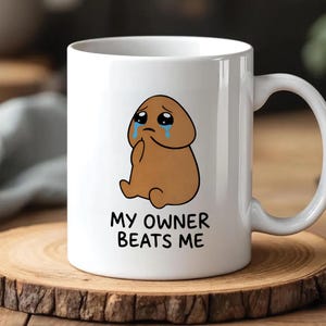 以下が含まれることがあります： 悲しそうな茶色のキャラクターと青い涙の漫画イラストが描かれた白いセラミックマグカップ。「MY OWNER BEATS ME」の文字がイラストの下に黒で印刷されています。マグカップには湾曲したハンドルが付いています。