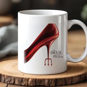 Puede incluir: Taza de cerámica blanca con un diseño de zapato de tacón alto rojo brillante, un tridente y el texto "The Devil Wears Prada". La taza tiene un asa curva y está sobre una superficie de madera.