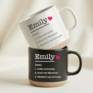 Puede incluir: Dos tazas de cerámica, una blanca y otra negra, apiladas una encima de la otra. Cada taza presenta el nombre "Emily" con un corazón, seguido de una definición que incluye "entusiasta del café" y "defensor de la siesta de fin de semana".