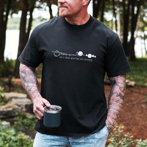 May include: Black t-shirt with white graphic design and the text "Life is about more than just surviving." The person is holding a dark blue travel mug. The background is a blurred outdoor scene with trees.