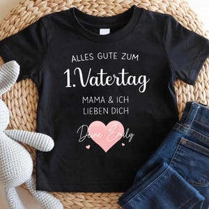 Puede incluir: Camiseta negra con letras blancas que dicen "Alles Gute Zum 1. Vatertag" y "Mama & Ich Lieben Dich". Un corazón rosa con el nombre "Deine Emily" está centrado debajo del texto. Vaqueros azul oscuro doblados junto a la camiseta.