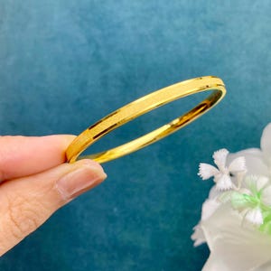 Pode incluir: Uma pulseira dourada com uma faixa central texturizada e bordas lisas. A pulseira &eacute; segurada por uma m&atilde;o contra um fundo azul, com flores brancas no canto. A joia tem um design simples e elegante.