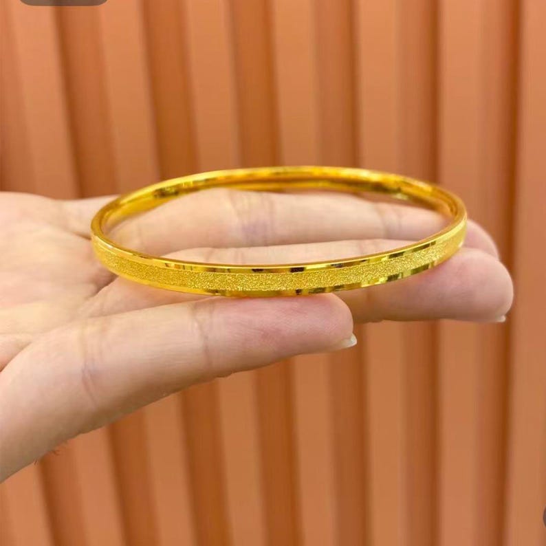 Pode incluir: Uma pulseira de ouro com uma faixa central texturizada. A pulseira &eacute; de cor dourada brilhante e parece ser feita de metal. A pulseira &eacute; redonda e lisa nas bordas.