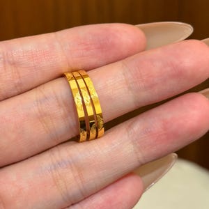 Puede incluir: Un anillo de oro con tres bandas, cada una con un diseño grabado repetido. El anillo es de color oro brillante y brillante y parece que se lleva en un dedo. El diseño grabado parece ser un patrón floral o de sol estilizado.