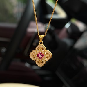 Puede incluir: Un colgante de oro con un diseño floral de cuatro lóbulos. El centro contiene cuatro pequeños rubíes rojos que rodean una piedra clara. Los pétalos están rellenos de filigrana dorada y bordeados por diminutas piedras claras en pavé.