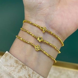 May include: Three gold bracelets are worn on a wrist. Each bracelet is made of small, faceted beads. One bracelet has a cross charm, another has a star, and the third has a four-leaf clover charm. The bracelets are a bright, shiny gold color.