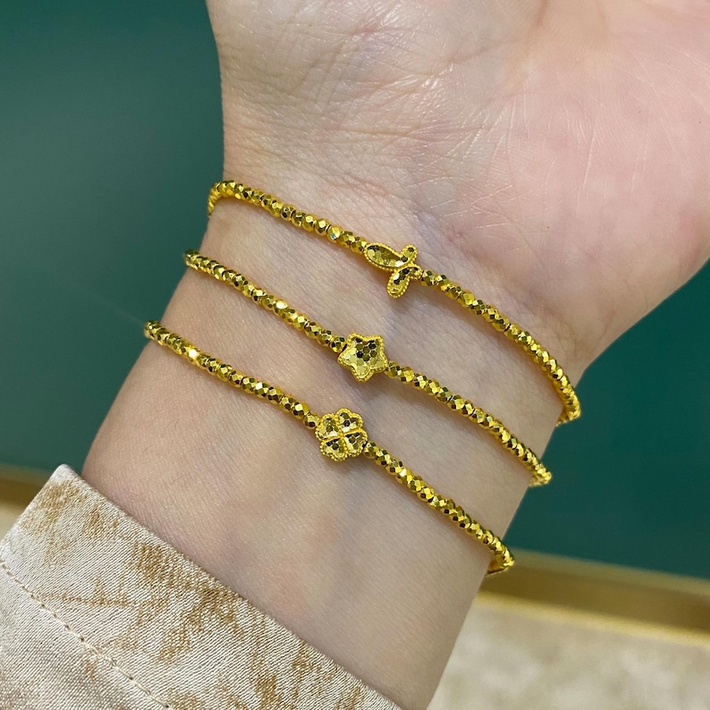 May include: Three gold bracelets are worn on a wrist. Each bracelet is made of small, faceted beads. One bracelet has a cross charm, another has a star, and the third has a four-leaf clover charm. The bracelets are a bright, shiny gold color.