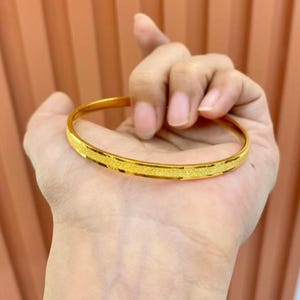 Pode incluir: Uma pulseira de ouro com uma faixa central texturizada. A pulseira &eacute; segurada em uma m&atilde;o, mostrando seu acabamento polido e design simples. A cor dourada d&aacute; &agrave; pulseira um visual cl&aacute;ssico e elegante.