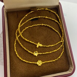 May include: Gold-colored beaded bracelets with charm accents, including a butterfly, star, and four-leaf clover, presented in a maroon jewelry box. The bracelets are displayed on a brown velvet-like surface.