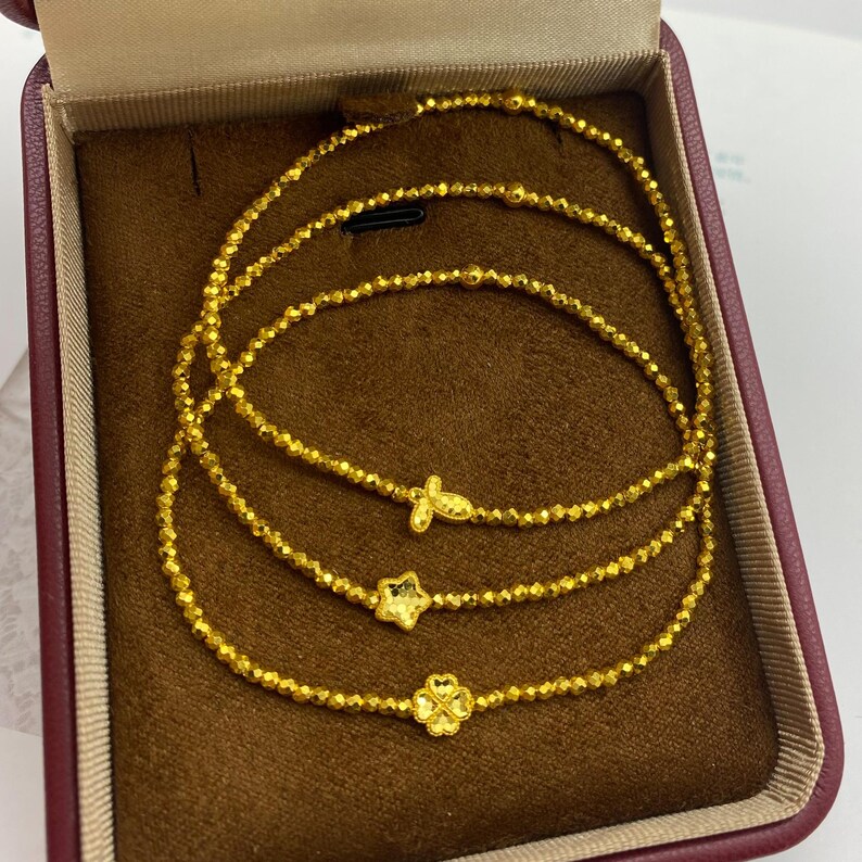 May include: Gold-colored beaded bracelets with charm accents, including a butterfly, star, and four-leaf clover, presented in a maroon jewelry box. The bracelets are displayed on a brown velvet-like surface.