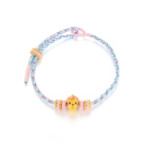 Puede incluir: Una pulsera trenzada con un patrón azul claro, rosa y morado. La pulsera presenta un dije dorado en el centro con un diseño de pollito de dibujos animados. Tiene cordones rosas ajustables y detalles dorados.