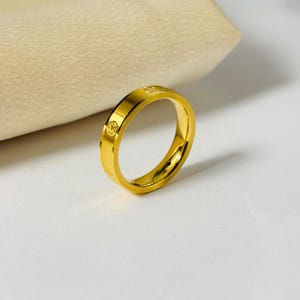 May include: A gold-colored ring with a smooth, polished finish. The ring features a series of small, decorative circular patterns around the band. The ring is set against a neutral background, highlighting its bright, reflective surface. A piece of jewelry.
