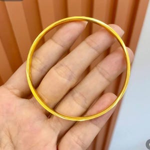 Pode incluir: Uma pulseira de argola dourada. A pulseira circular &eacute; lisa e brilhante, refletindo a luz. A joia &eacute; segurada em uma m&atilde;o, mostrando seu design simples e elegante. A pulseira &eacute; uma pe&ccedil;a cl&aacute;ssica de joalheria.
