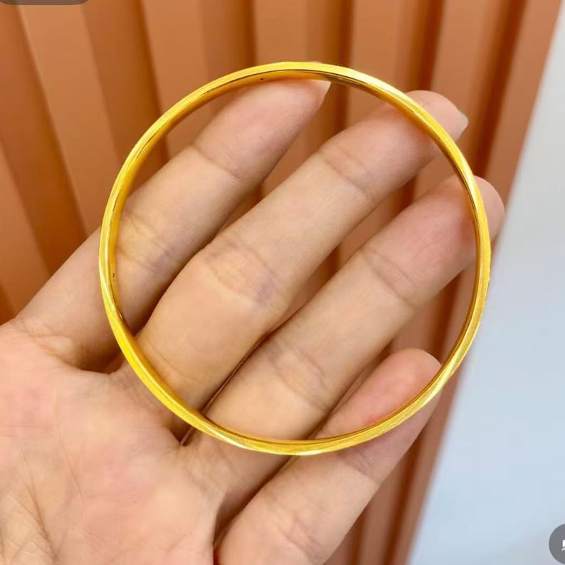 Pode incluir: Uma pulseira de argola dourada. A pulseira circular &eacute; lisa e brilhante, refletindo a luz. A joia &eacute; segurada em uma m&atilde;o, mostrando seu design simples e elegante. A pulseira &eacute; uma pe&ccedil;a cl&aacute;ssica de joalheria.