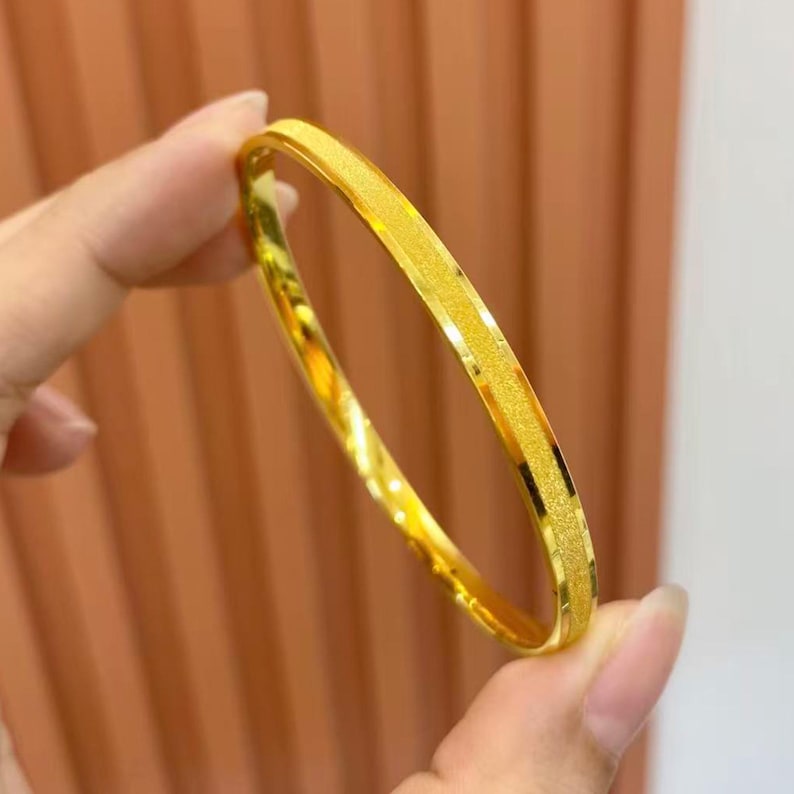 Pode incluir: Uma pulseira de ouro com uma faixa central texturizada e brilhante. A pulseira &eacute; segurada por uma m&atilde;o, mostrando sua apar&ecirc;ncia brilhante e brilhante. A joia tem um design simples e elegante.