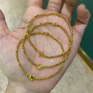 May include: Three gold bracelets with a beaded design are displayed. Each bracelet features a different charm: a star, a four-leaf clover, and a butterfly. The bracelets are made of small, faceted beads that catch the light.