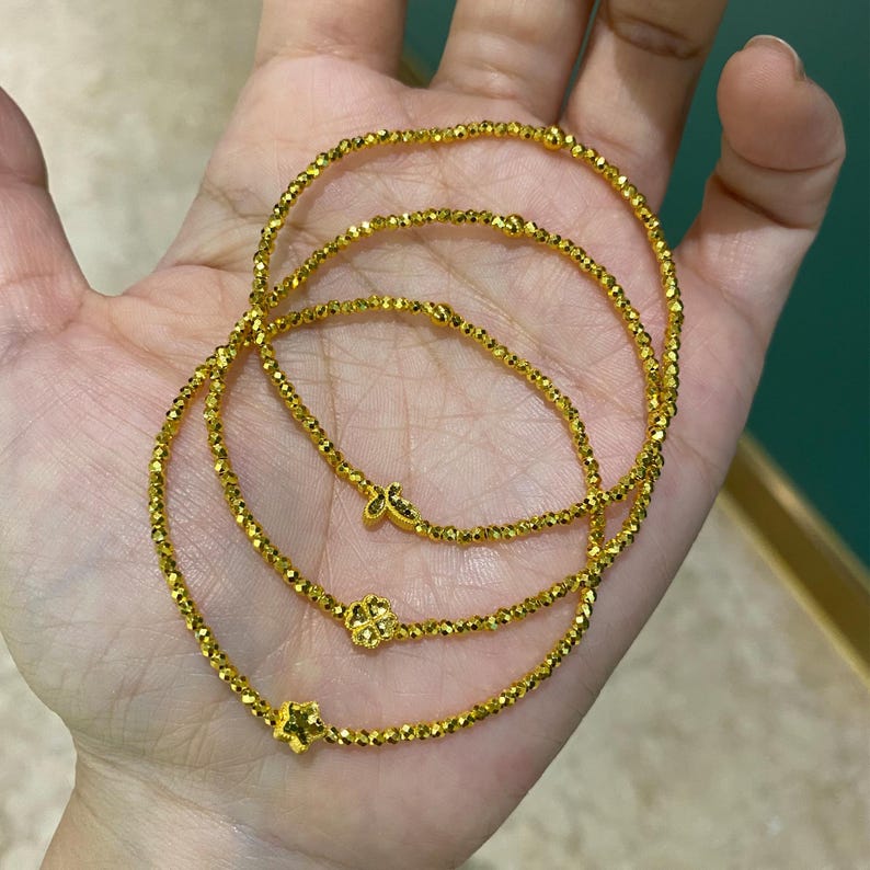 May include: Three gold bracelets with a beaded design are displayed. Each bracelet features a different charm: a star, a four-leaf clover, and a butterfly. The bracelets are made of small, faceted beads that catch the light.