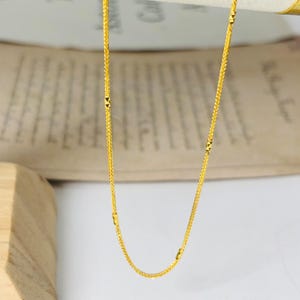 May include: A delicate gold chain necklace with a woven design. The necklace features small, decorative accents and hangs against a neutral background. The chain is a bright, shiny gold colour.