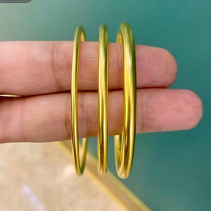 May include: Three gold-colored bangles of varying thicknesses are displayed. The bangles are smooth and shiny, reflecting light. The background is a teal color. The bangles are likely made of metal.