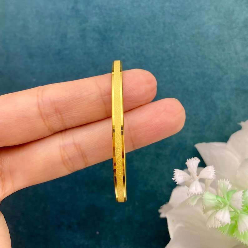 Pode incluir: Uma pulseira dourada com superf&iacute;cie texturizada e formato retangular. A pulseira &eacute; segurada em uma m&atilde;o contra um fundo azul-petr&oacute;leo com flores brancas.