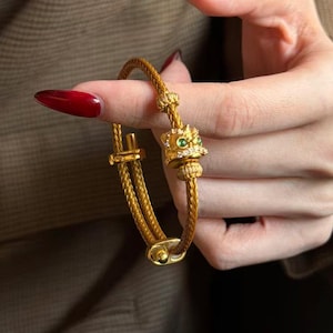 May include: A gold-toned bracelet featuring a braided cable band and a central charm shaped like a stylised lion head. The charm is accented with small clear stones and two green circular stones for eyes, with a sliding clasp mechanism at the base.