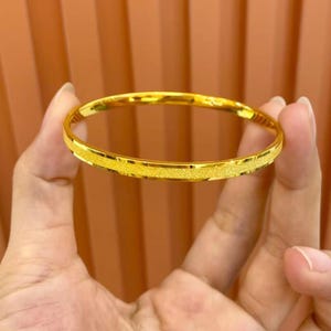 Pode incluir: Uma pulseira dourada com uma faixa texturizada e brilhante. A pulseira &eacute; segurada em frente a um fundo castanho. A pulseira &eacute; uma pe&ccedil;a de joalharia simples e elegante.