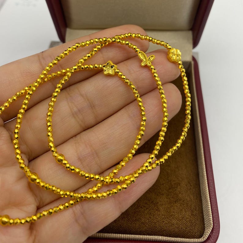 May include: A delicate gold necklace with three strands of small, faceted beads. The necklace features a star, butterfly, and flower charm. The jewelry is displayed inside a red jewelry box.