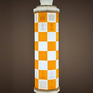 May include: A tall, cylindrical lamp with a white and orange checkered pattern. The lamp features chess piece designs and a crown-shaped top. The base and top are white, and the lamp is illuminated, creating a warm glow.