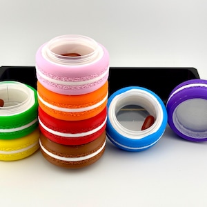 May include: A collection of colorful, macaron-shaped containers in various colors, including pink, orange, red, brown, blue, green, yellow, and purple. Each container has a white accent and a clear lid. The containers are arranged on a white surface.