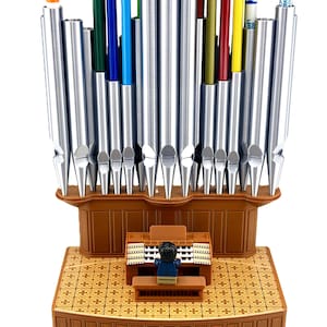 May include: A brown plastic organ-shaped pencil holder featuring a miniature figure seated at a console. The structure includes vertical silver metallic tubes of varying heights that serve as slots for colorful pencils and markers.