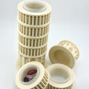 May include: A beige, miniature replica of the Leaning Tower of Pisa, with a clear plastic container inside. Several detached sections are visible, with one containing pills. The tower is a decorative item.