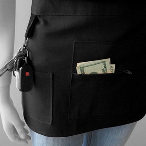 Op de afbeelding: Een zwarte schort met meerdere zakken, waaronder een zak met rits. Er zitten dollarbiljetten in de zak en een sleutelbos aan een ring aan de zijkant.