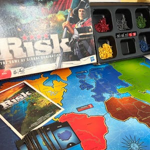 Könnte beinhalten: Das Brettspiel Risk: The Game of Global Domination mit Spielbrett, Karten, Würfeln und Spielfiguren in Rot, Grau, Grün, Gelb und Blau. Das Box-Art zeigt einen Soldaten mit einer Flagge. Das Spiel ist für Kinder ab 10 Jahren geeignet.