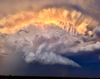 Könnte beinhalten: Eine große, dramatische Gewitterwolke mit einem leuchtend orangenen Schein an der Spitze und einer dunklen, wirbelnden Basis. Die Wolke wird vom untergehenden Sonnenlicht beleuchtet, was einen starken Kontrast zwischen den hellen und dunklen Bereichen erzeugt.