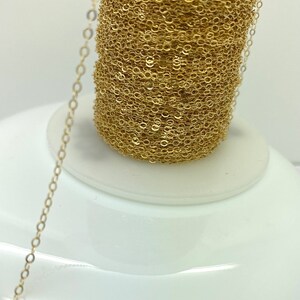 May include: A spool of delicate gold-colored chain, with a fine, oval link design. The chain is tightly wound on a white base, with a length of the chain extending out. The chain is likely used for jewelry making.