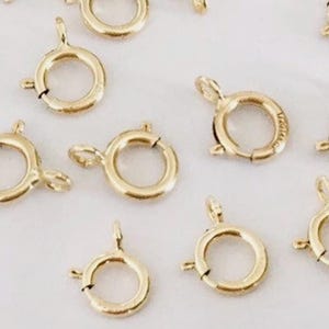 May include: Close-up of several gold-colored spring ring clasps. These jewelry findings are round with a small lever to open and close the clasp. The clasps are used to secure necklaces, bracelets, and other jewelry.
