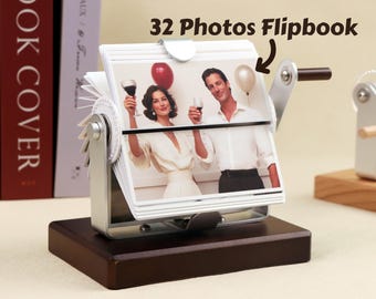 May include: A white photo flipbook with a wooden base. The flipbook holds 32 photos and has a crank to turn the pages. The text "32 Photos Flipbook" is written on the image.