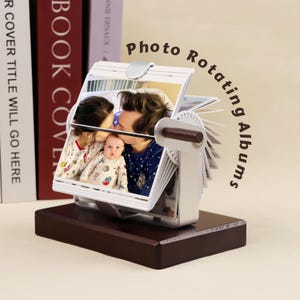 May include: A white photo album with a wooden handle rotates to display photos of a family. The album is on a dark brown wooden base. The text "Photo Rotating Albums" is written in brown on a white background.