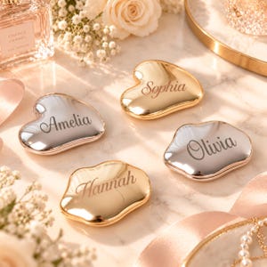 May include: Four heart-shaped compact mirrors with metallic finishes in gold and silver. Each mirror is engraved with a unique name: Amelia, Sophia, Olivia, and Hannah. The mirrors have a smooth, reflective surface and irregular, organic edges.
