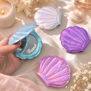 Pode incluir: Quatro espelhos compactos em forma de concha com acabamento iridescente. Um espelho azul-claro está aberto, revelando uma superfície de vidro circular com o nome Hannah impresso em letra branca. Os outros três espelhos estão fechados em branco, roxo e rosa.