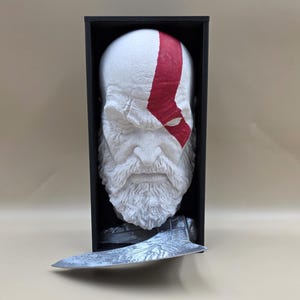 Puede incluir: Busto blanco de un hombre barbudo con una franja roja en la cara, exhibido en una caja negra. Un hacha plateada se encuentra debajo del busto. El busto tiene una superficie detallada y texturizada.