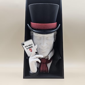Può includere: Scultura di un uomo con cilindro e abito che tiene in mano una carta da gioco con la scritta "IT'S ONLY A GAME". Il cappello è nero con una fascia rossa e il volto dell'uomo è di colore chiaro. La scultura è in una cornice nera.