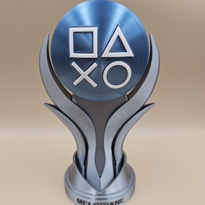 Op de afbeelding: Een zilveren trofee met een blauwe bol erop. De bol heeft de PlayStation-knopsymbolen erop. De basis van de trofee zegt "MELODIANE".