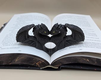 Può includere: Un fermapagina a forma di drago nero, a forma di due draghi che si fronteggiano, con un'apertura a forma di cuore. I draghi sono posizionati su un libro aperto, con testo visibile sulle pagine. I draghi hanno occhi gialli.